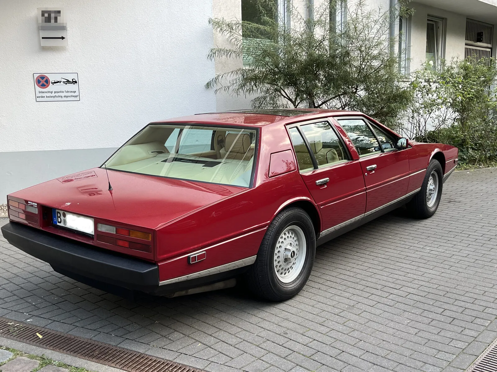 Roter Aston Martin Lagonda von schräg hinten, geparkt vor einem weißen Wohnhaus