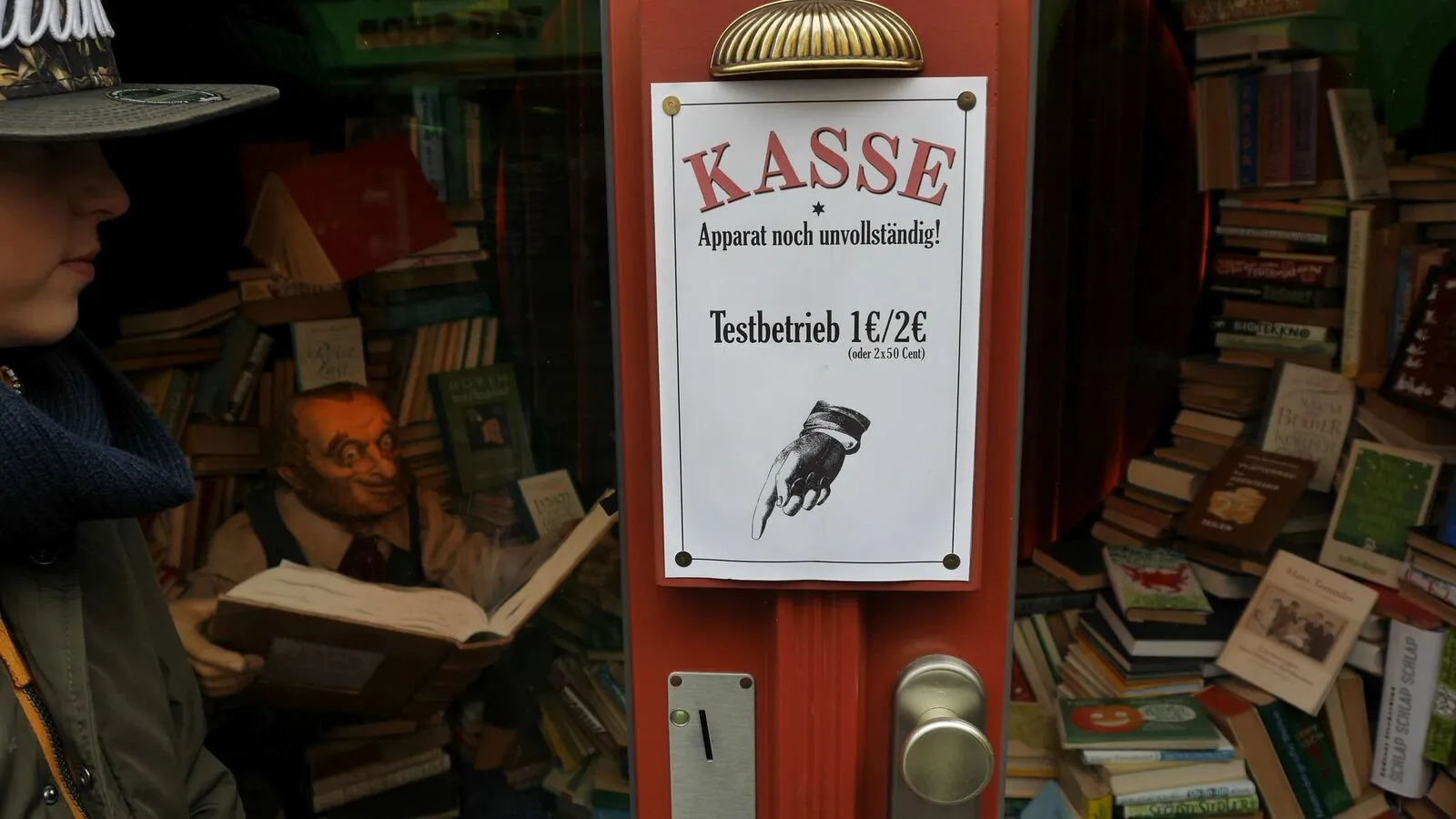 Rote Ladentür mit dem Aushang „Kasse – Apparat noch unvollständig! Testbetrieb 1€/2€“ und Münzschlitz, dahinter ein Schaufenster voller Bücher mit einer lesenden Figur.