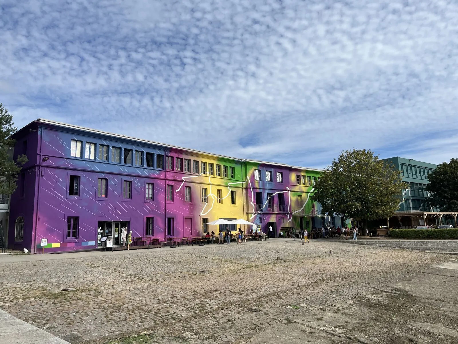 Dreistöckiges Gebäude mit Fassade im Farbverlauf von Lila über Gelb nach Grün, davor ein weiter Platz mit Kopfsteinpflaster und Menschen unter Sonnenschirmen.